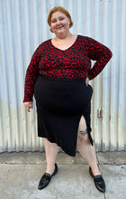 Load image into Gallery viewer, Additional full-body front view of a size 26 Lane Bryant black stretchy midi skirt with convertible high side slit styled with a red & black leopard bodysuit and black loafers on a size 22/24 model. The photo is taken outside in natural lighting.