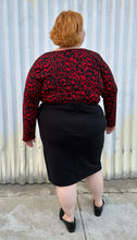 Load image into Gallery viewer, Full-body back view of a size 26 Lane Bryant black stretchy midi skirt with convertible high side slit styled with a red & black leopard bodysuit and black loafers on a size 22/24 model. The photo is taken outside in natural lighting.