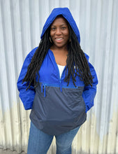 Load image into Gallery viewer, Front view of a size XL Harriton cobalt and navy blue windbreaker styled unzipped with the hood up with jeans on a size 14/16 model. The photo is taken outside in natural lighting.