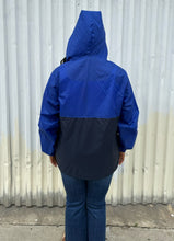 Load image into Gallery viewer, Back view of a size XL Harriton cobalt and navy blue windbreaker styled unzipped with the hood up with jeans on a size 14/16 model. The photo is taken outside in natural lighting.