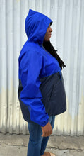 Load image into Gallery viewer, Side view of a size XL Harriton cobalt and navy blue windbreaker styled unzipped with the hood up with jeans on a size 14/16 model. The photo is taken outside in natural lighting.