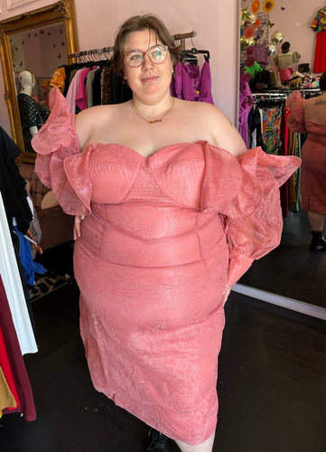 Front view of a size 24 ASOS peachy pink lace sheath dress with a corset-style bodice and oversized ruffle bust and sleeves with puff sleeves on a size 24 model. The photo is taken inside in front of a window, for a combination of natural and overhead lighting.