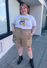 Load image into Gallery viewer, Full-body front view of a size XL &quot;Evel Pie&quot; Evel Knievel pizza t-shirt styled tucked into brown shorts on a size 22/24 model. The photo was taken outside in natural lighting.
