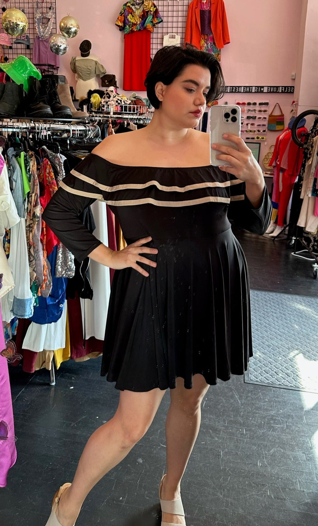 Full-body front view of a size 3X Rue 107 black and tan ruffled neckline off-the-shoulder long sleeve mini dress styled with tan heels on a size 14/16 model. The photo was taken inside under fluorescent lighting.