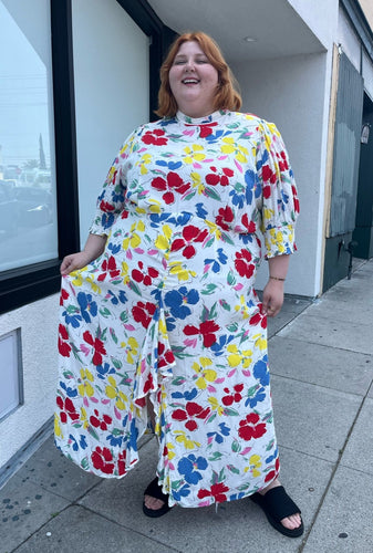Full-body front view of a size 28/30W RIXO x Target collab white and rainbow floral high-necked maxi dress with a ruffled front slit on a size 22/24 model. The photo was taken outside in natural lighting.