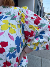 Load image into Gallery viewer, Close up view of the sketched rainbow floral pattern of a size 28/30W RIXO x Target collab white and rainbow floral high-necked maxi dress with a ruffled front slit on a size 22/24 model. The photo was taken outside in natural lighting.
