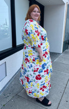 Load image into Gallery viewer, Full-body side view of a size 28/30W RIXO x Target collab white and rainbow floral high-necked maxi dress with a ruffled front slit on a size 22/24 model. The photo was taken outside in natural lighting.
