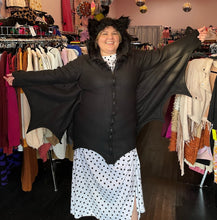 Load image into Gallery viewer, Front view of a size 1/2X Leg Avenue black bat costume hoodie with bat wing, connected sleeves and fuzzy ears on the hood, styled zipped closed over a white and black polka dot dress on a size 14/16 model.
