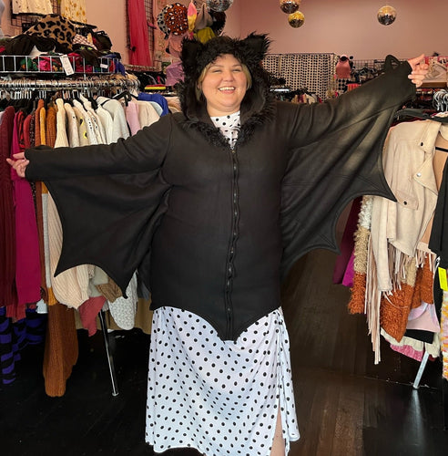 Front view of a size 1/2X Leg Avenue black bat costume hoodie with bat wing, connected sleeves and fuzzy ears on the hood, styled zipped closed over a white and black polka dot dress on a size 14/16 model.