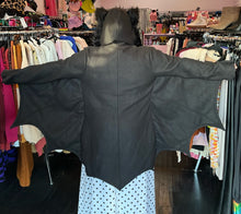 Load image into Gallery viewer, Back view of a size 1/2X Leg Avenue black bat costume hoodie with bat wing, connected sleeves and fuzzy ears on the hood, styled over a white and black polka dot dress on a size 14/16 model.
