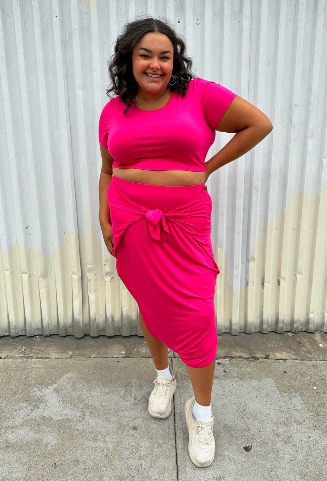 Full-body front view of a size 2 Fashion to Figure two piece hot pink cropped tee and tie-detail maxi skirt set styled with white sneakers on a size 18/20 model. The photo is taken outside in natural lighting.