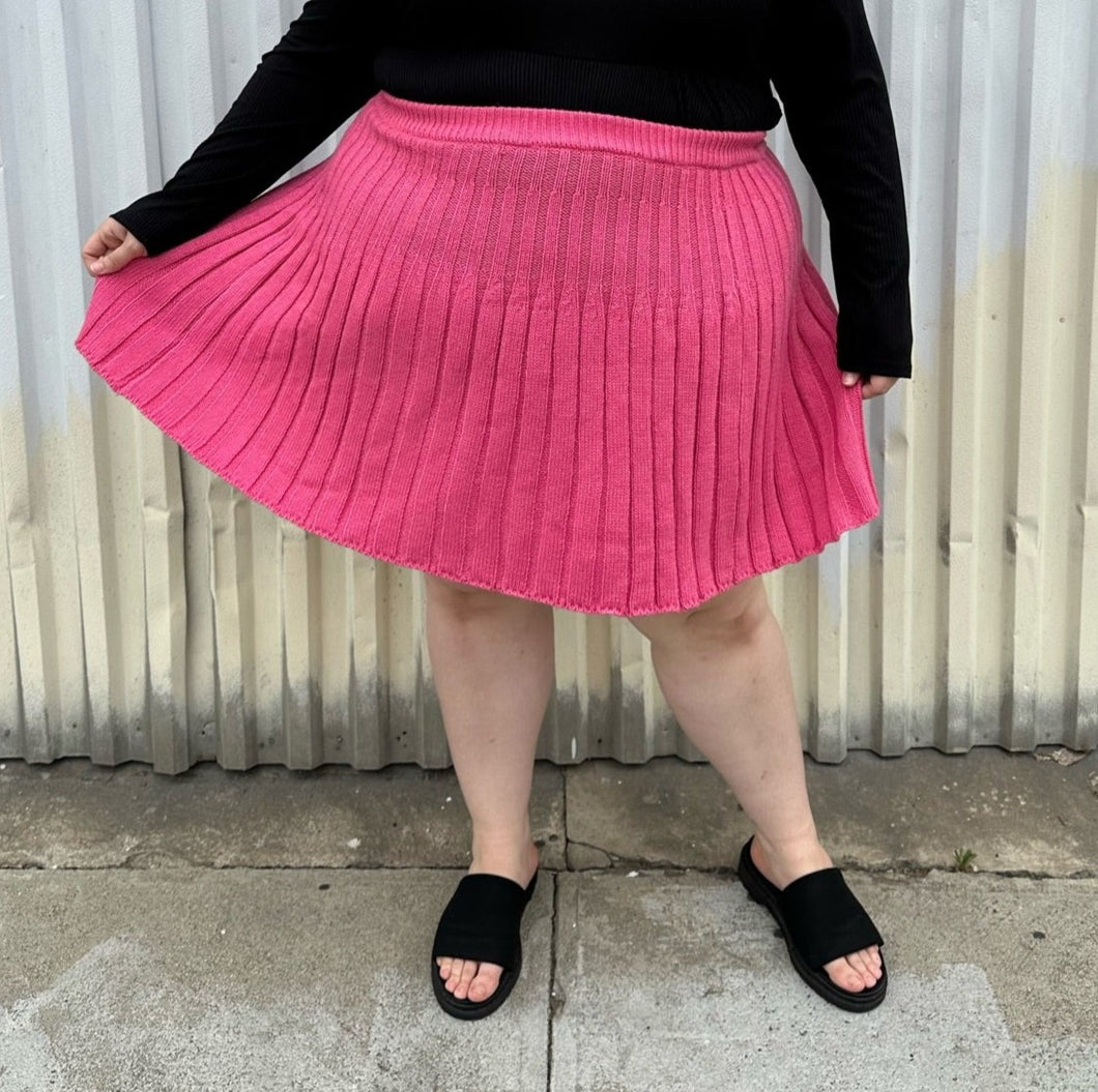 Front view of a size 22/24 Eloquii bubblegum pink mini circle skirt made of sweater material styled with a long sleeve black shirt and black slides on a size 22/24 model. The photo is taken outside in natural lighting.