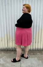 Load image into Gallery viewer, Full-body side view of a size 22/24 Eloquii bubblegum pink mini circle skirt made of sweater material styled with a long sleeve black shirt and black slides on a size 22/24 model. The photo is taken outside in natural lighting.
