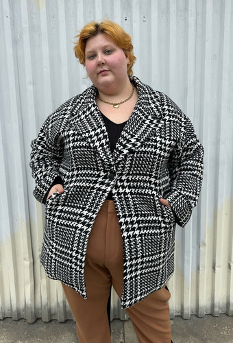 Front view of a size 22/24 Eloquii black and white houndstooth peacoat styled closed with its single-button closure over a black long sleeve and brown trousers on a size 22/24 model. The photo is taken outside in natural lighting.