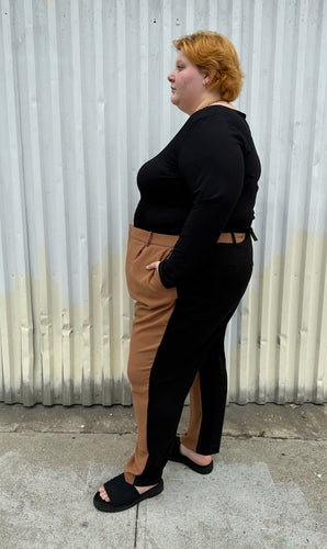 Full-body side view of a pair of size 28 Eloquii half brown, half black trousers styled with a black long sleeve and black slides on a size 22/24 model. The photo is taken outside in natural lighting.