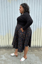 Load image into Gallery viewer, Full-body side view of a size 4X (fits like 2X) Banned Retro black maxi skirt with orange, purple, and white mixed Halloween pattern featuring cats with witch hats styled with a black long sleeve and white mules on a size 18/20 model. The photo was taken outside in natural lighitng.
