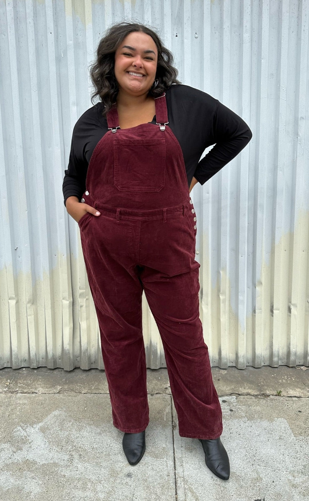 Full-body front view of a pair of size 26 (fits like 18/20) Ulla Popken maroon corduroy overalls styled over a black long sleeve and boots on a size 18/20 model. The photo is taken outside in natural lighting.