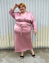 Load image into Gallery viewer, Full-body front view of a size 22/24 Eloquii blush pink silky faux wrap maxi dress with a belt styled with black kitten heels on a size 22/24 model. The photo was taken outside in natural lighting.
