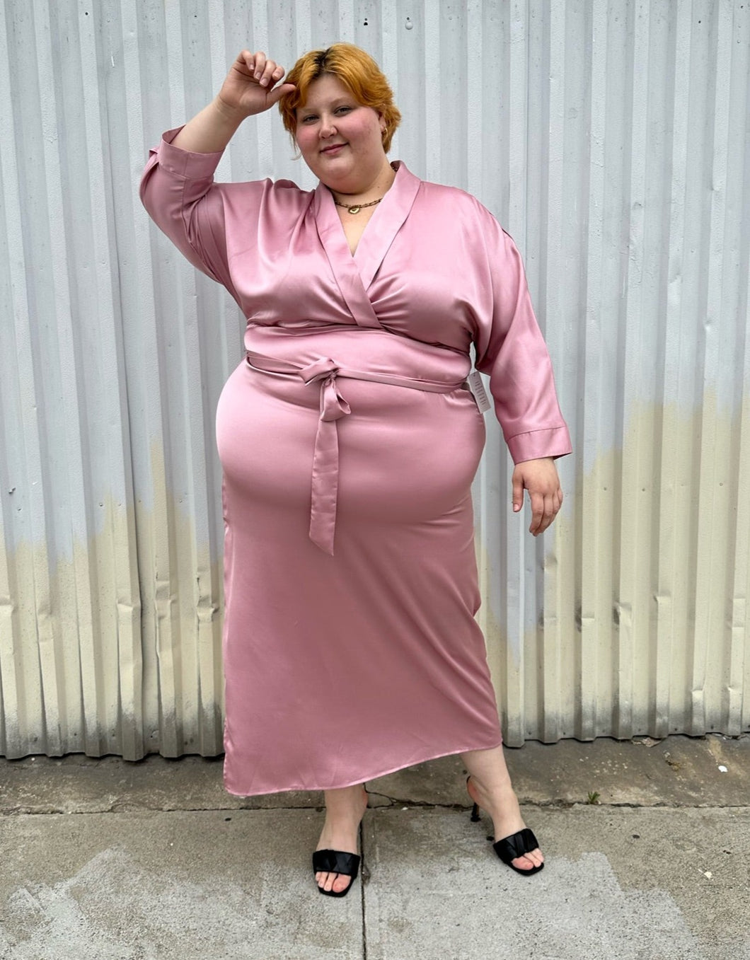 Full-body front view of a size 22/24 Eloquii blush pink silky faux wrap maxi dress with a belt styled with black kitten heels on a size 22/24 model. The photo was taken outside in natural lighting.