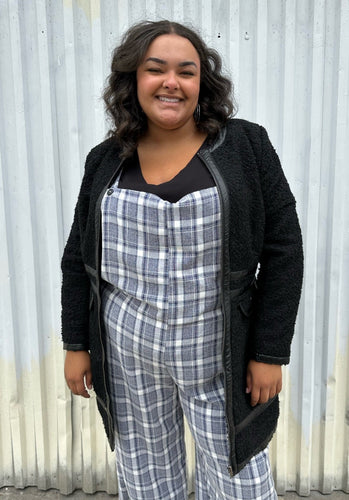 Front view of a size 18 City Chic black collarless longline zip-up jacket with pleather accents styled open over a blue & white plaid jumpsuit on a size 18/20 model. The photo was taken outside in natural lighting.