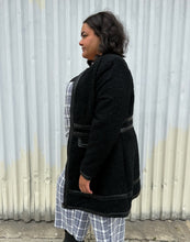Load image into Gallery viewer, Side view of a size 18 City Chic black collarless longline zip-up jacket with pleather accents styled open over a blue &amp; white plaid jumpsuit on a size 18/20 model. The photo was taken outside in natural lighting.

