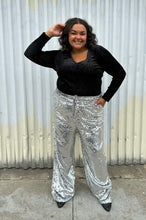 Load image into Gallery viewer, Full-body front view of a size 18/20 Eloquii black velvet long sleeve v-neck top with silver metallic threading throughout styled tucked into silver sequin trousers on a size 18/20 model. The photo was taken outside in natural lighting.

