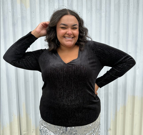Front view of a size 18/20 Eloquii black velvet long sleeve v-neck top with silver metallic threading throughout styled over silver sequin trousers on a size 18/20 model. The photo was taken outside in natural lighting.