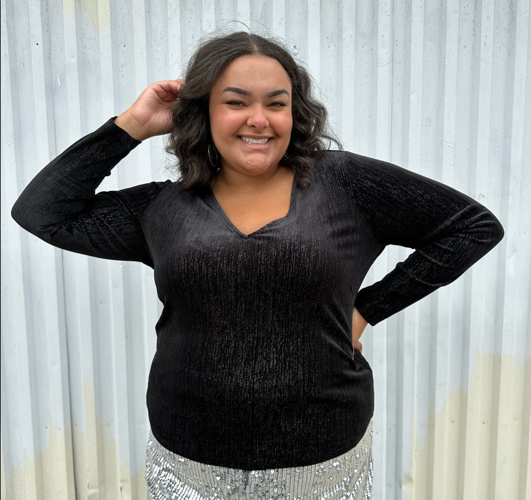 Front view of a size 18/20 Eloquii black velvet long sleeve v-neck top with silver metallic threading throughout styled over silver sequin trousers on a size 18/20 model. The photo was taken outside in natural lighting.