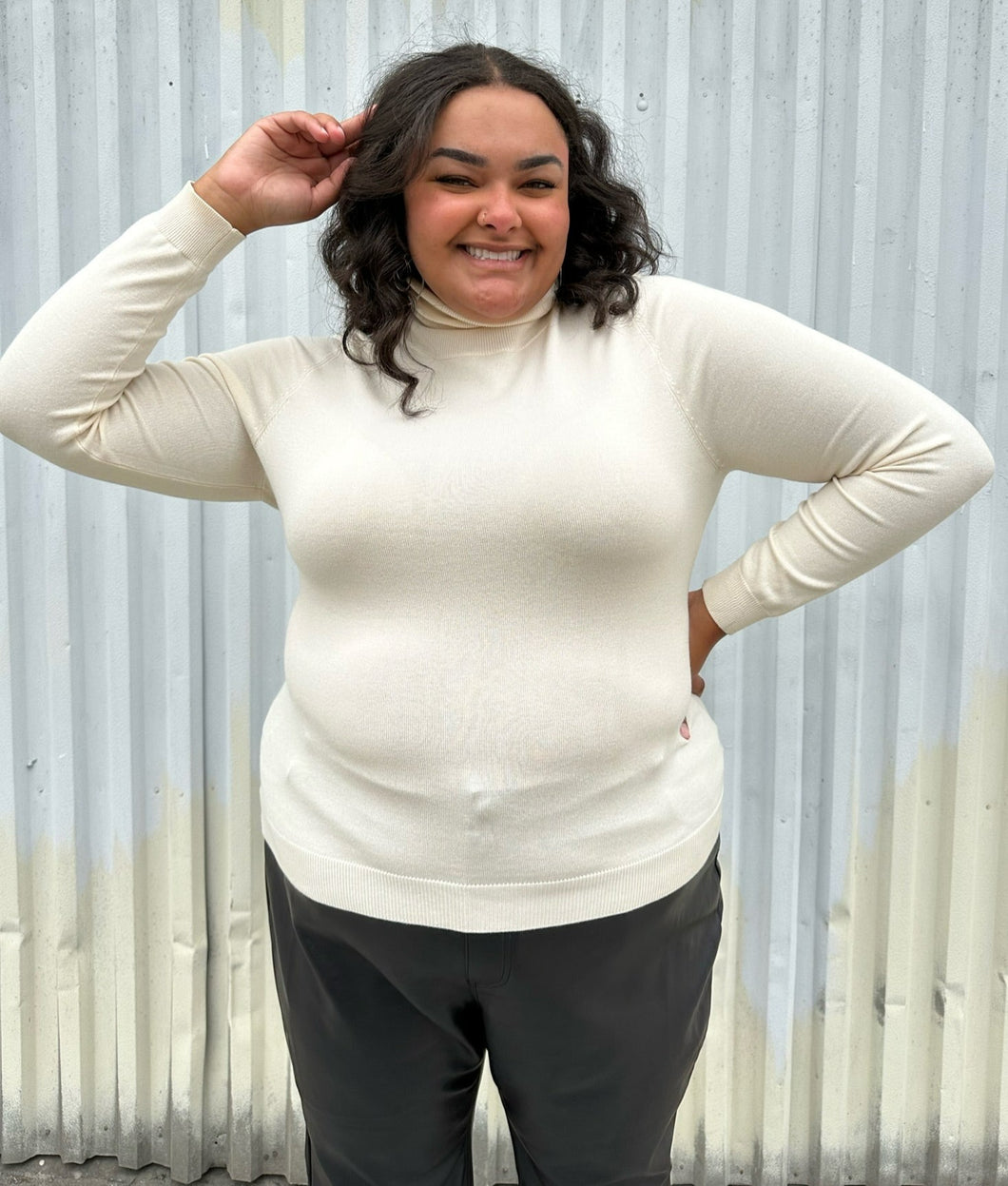 Front view of a size 2 Fashion to Figure cream turtleneck sweater styled with black trousers on a size 18/20 model. The photo was taken outside in natural lighting.