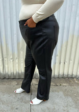 Load image into Gallery viewer, Side view of a size 24 (fits like 20/22) Eloquii black pleather straight leg pants styled with a cream turtleneck and white mules on a size 18/20 model. The photo was taken outside in natural lighting.
