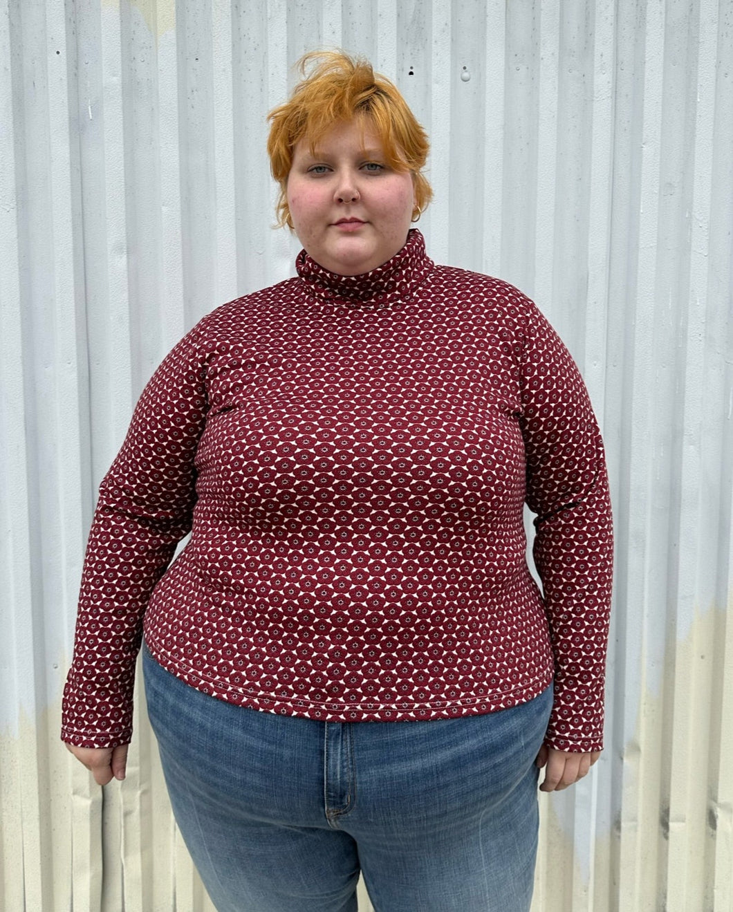 Front view of a size 4 Fashion to Figure x Gabi Fresh maroon & off-white 70s floral turtleneck top styled over medium wash denim on a size 22/24 model. The photo is taken outside in natural lighting.