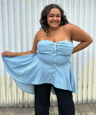 Front view of a size 2 Fashion to Figure chambray strapless button-up high-low blouse styled over dark wash denim on a size 18/20 model. The photo was taken outside in natural lighting.
