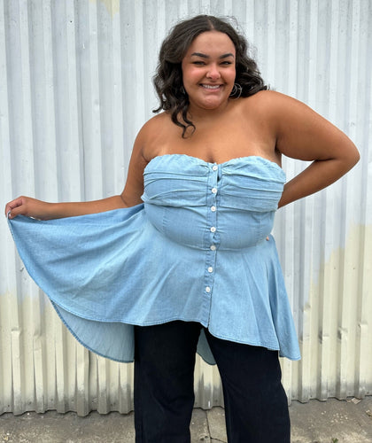 Front view of a size 2 Fashion to Figure chambray strapless button-up high-low blouse styled over dark wash denim on a size 18/20 model. The photo was taken outside in natural lighting.