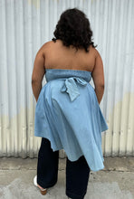 Load image into Gallery viewer, Full-body back view of a size 2 Fashion to Figure chambray strapless button-up high-low blouse styled over dark wash denim on a size 18/20 model. The photo was taken outside in natural lighting.
