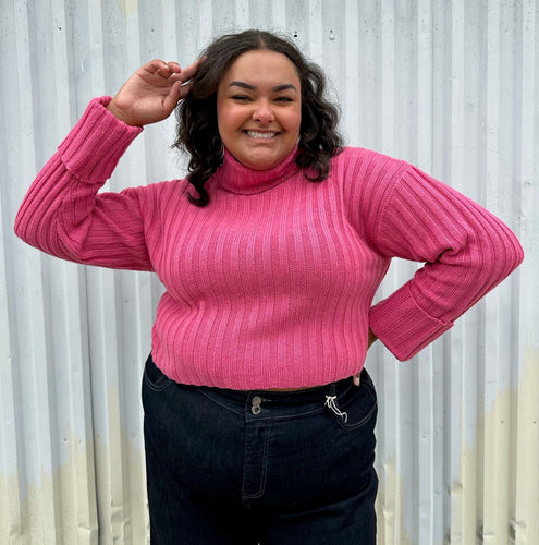 Front view of a size 18/20 Eloquii bubblegum pink ribbed turtleneck cropped sweater styled over a dark wash denim trouser on a size 18/20 model The photo was taken outside in natural lighting.