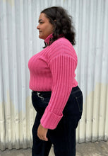 Load image into Gallery viewer, Side view of a size 18/20 Eloquii bubblegum pink ribbed turtleneck cropped sweater styled over a dark wash denim trouser on a size 18/20 model The photo was taken outside in natural lighting.
