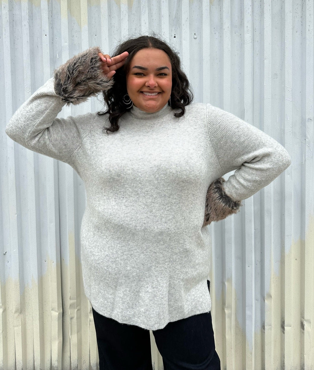 Front view of a size 18/20 Lane Bryant x Girl with Curves collab gray ribbed longline turtlneck sweater with brown faux fur cuffs styled with dark wash denim on a size 18/20 model. The photo was taken outside in natural lighting.