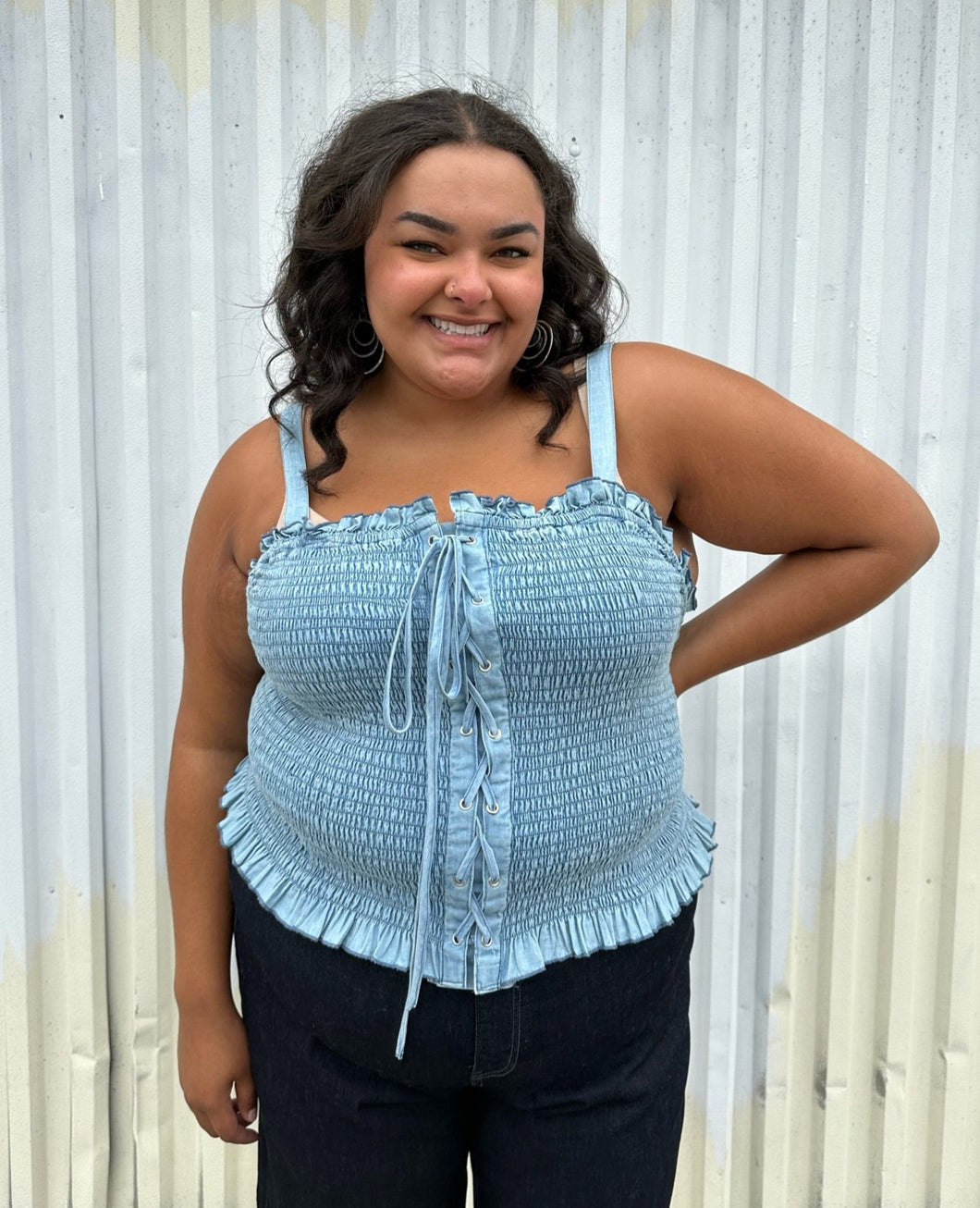 Front view of a size 2 Fashion to Figure chambray all-over smocked tank with a corset-style lace up front styled over dark wash denim on a size 18/20 model. The photo was taken outside in natural lighting.