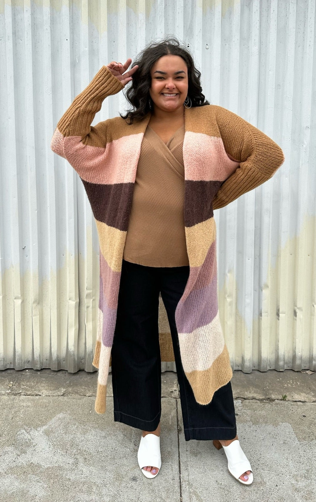 Full-body front view of a size XL (fits comfy up to 22) Free People brown, cream, pink, and purple striped longline open knit cardigan sweater styled open over a light brown top. dark wash denim, and white mules on a size 18/20 model. The photo was taken outside in natural lighting.