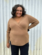 Load image into Gallery viewer, Front view of a size 18/20 Eloquii light brown ribbed faux wrap sweater styled over dark wash denim on a size 18/20 model. The photo was taken outside in natural lighting.
