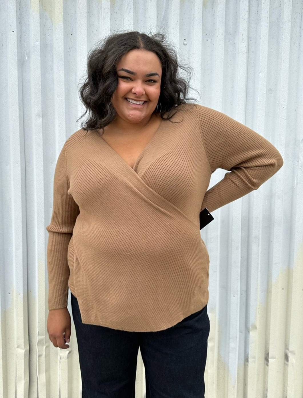 Front view of a size 18/20 Eloquii light brown ribbed faux wrap sweater styled over dark wash denim on a size 18/20 model. The photo was taken outside in natural lighting.