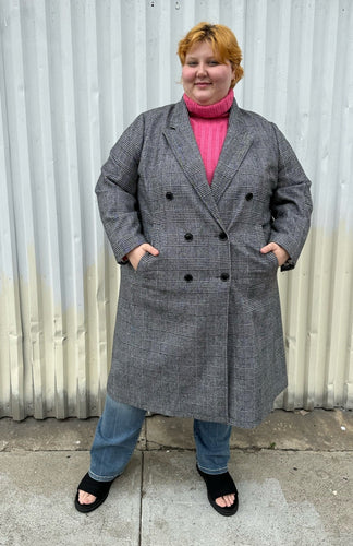 Full-body front view of a size 3X Nilli Lotan x Target black and white gingham double breasted longline peacoat styled closed over a pink sweater, medium wash denim, and black slides on a size 22/24 model. The photo was taken outside in natural lighting.
