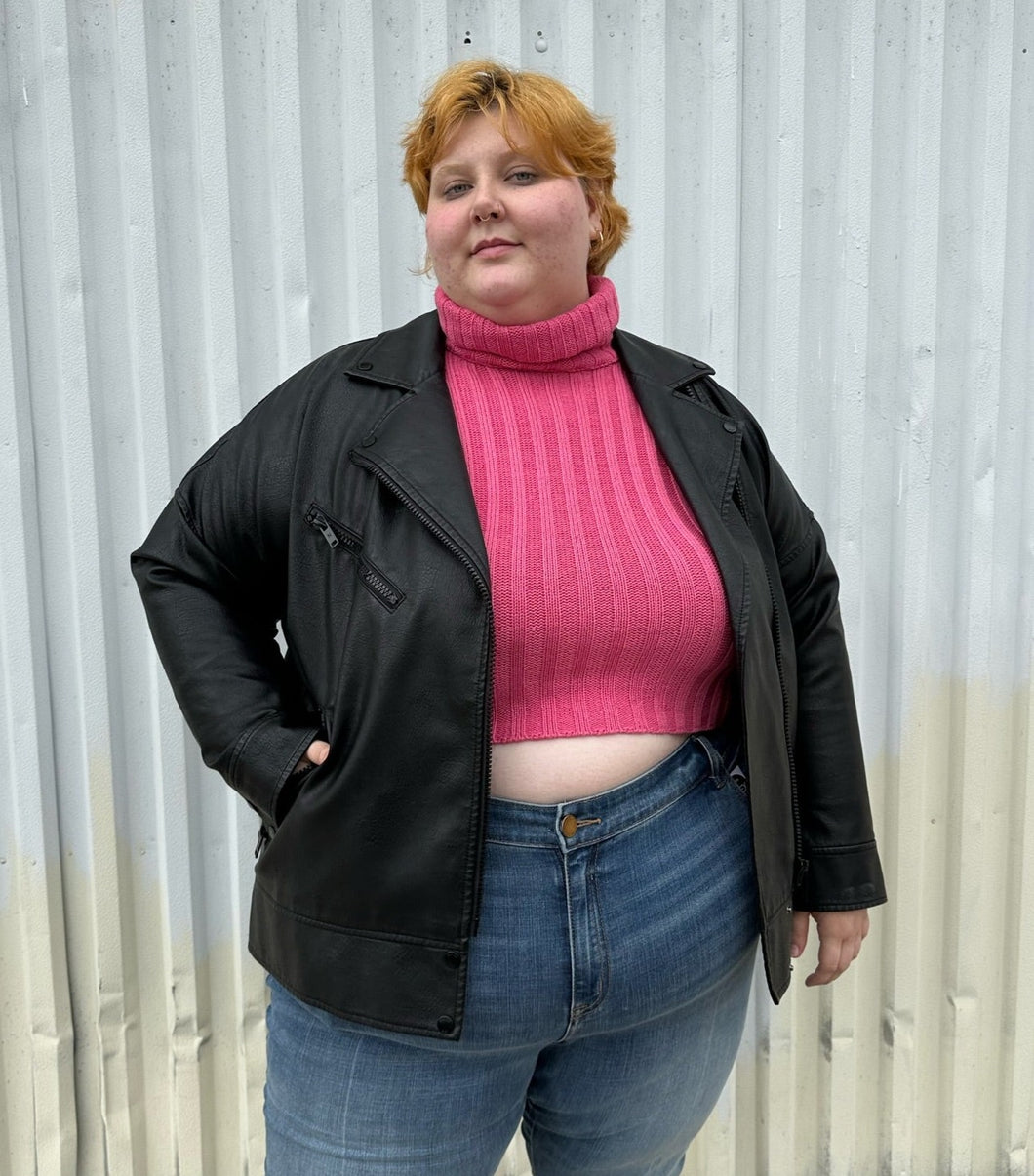 Front view of a size 48 Noisy May black faux leather moto jacket styled open over a pink cropped turtleneck and medium wash denim on a size 22/24 model. The photo was taken outside in natural lighting.