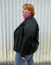 Load image into Gallery viewer, Side view of a size 48 Noisy May black faux leather moto jacket styled open over a pink cropped turtleneck and medium wash denim on a size 22/24 model. The photo was taken outside in natural lighting.
