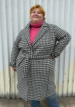 Load image into Gallery viewer, Front view of a size 22 Vero Moda black and white houndstooth plaid belted peacoat styled tied closed over a pink turtleneck and medium wash denim on a size 22/24 model. The photo was taken outside in natural lighting.
