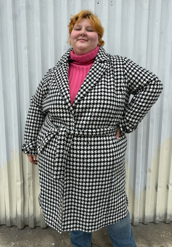 Front view of a size 22 Vero Moda black and white houndstooth plaid belted peacoat styled tied closed over a pink turtleneck and medium wash denim on a size 22/24 model. The photo was taken outside in natural lighting.