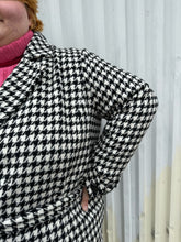 Load image into Gallery viewer, Close up view of the houndstooth pattern of a size 22 Vero Moda black and white houndstooth plaid belted peacoat styled tied closed over a pink turtleneck on a size 22/24 model. The photo was taken outside in natural lighting.
