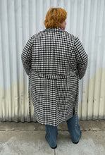 Load image into Gallery viewer, Full-body back view of a size 22 Vero Moda black and white houndstooth plaid belted peacoat styled tied closed over a pink turtleneck and medium wash denim on a size 22/24 model. The photo was taken outside in natural lighting.
