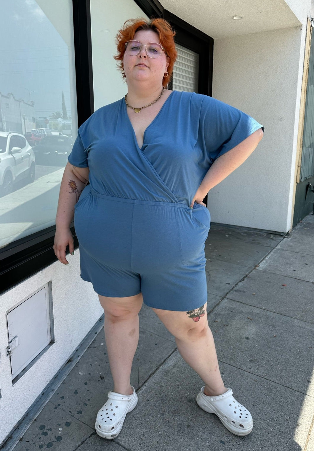 Full-body front view of a size L Universal Standard dusty blue stretchy romper with pockets styled with white platform sandals on a size 22/24 model. The photo was taken outside in natural lighting.