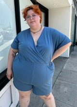 Load image into Gallery viewer, Front view of a size L Universal Standard dusty blue stretchy romper with pockets styled with white platform sandals on a size 22/24 model. The photo was taken outside in natural lighting.
